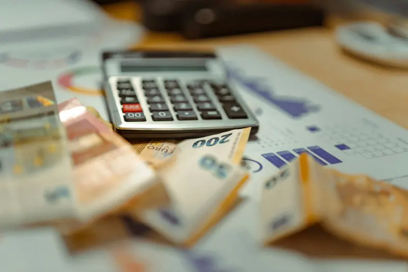 A calculator resting on top of a stack of euro banknotes and a printed budget spreadsheet